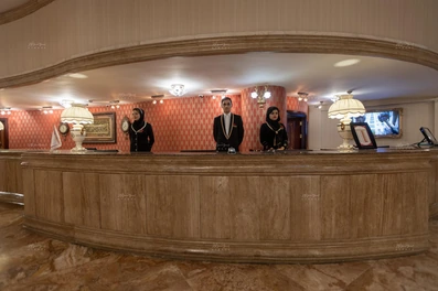 دو خانم و یک آقا با لباس فرم سورمه ای رنگ پشت پیشخان پذیرش در هتل گلدن پالاس ایستاده اند. پیشخان با تزئینات کلاسیک چوبی به شکل خمیده ساخته شده است.