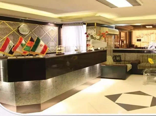 پذیرش هتل آپارتمان کوروش مشهد با نمای دیوارهای سنگی طوسی و سرامیکهای سفید در کف هتل و پرچمهای کشورهای مختلف